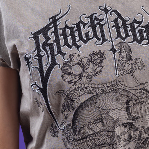 A close-up of the Black Decay Embroidered Female T-shirt, showing gothic black text, a rose, a snake, and detailed skull embroidery on gray acid-wash fabric. Part of a woman's arm and torso appear against a purple background.