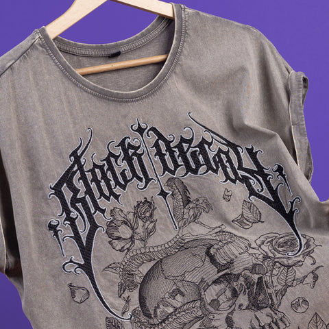 The Black Decay Embroidered Female T-shirt showcases bold gothic black text and detailed floral and ram’s skull embroidery on gray acid-wash fabric, displayed on a wooden hanger against a purple backdrop—a striking memento mori piece.