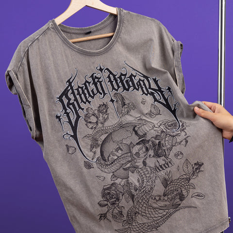 A hand holds the Black Decay Embroidered Female T-shirt on a wooden hanger, showcasing a gothic Angel Rebel logo and a detailed skull, rose, and snake illustration against a purple acid-wash background.