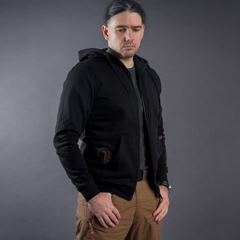 A man with long dark hair wears the Skull & Scolopendras Limited Edition Embroidered Hoodie, a gray t-shirt, and brown pants, standing against a plain gray background and looking down with a neutral expression.