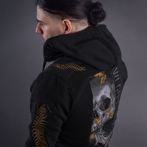 A person with dark hair in a ponytail, wearing the Skull & Scolopendras Limited Edition Embroidered Hoodie featuring gold centipede designs and a large skull graphic on the back, faces away against a dark gray background.