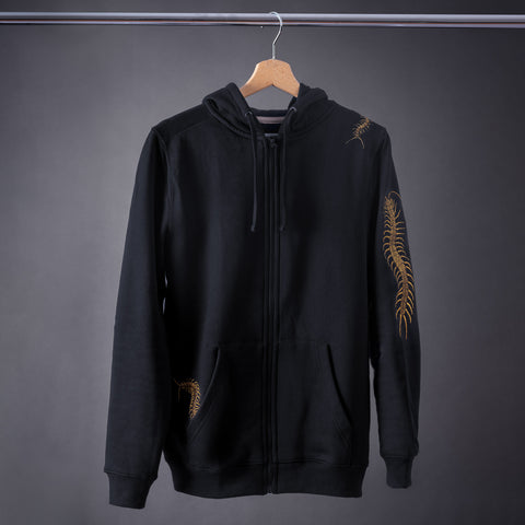 The Skull & Scolopendras Limited Edition Embroidered Hoodie is a black zip-up with golden centipede designs on the shoulder, sleeve, and pocket, shown hanging on a wooden hanger against a gray background.
