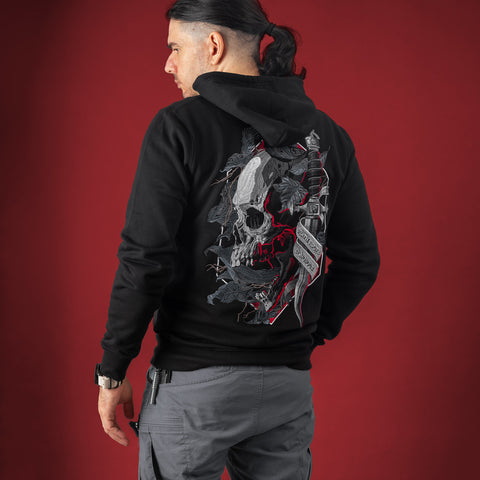 A man with dark hair in a ponytail dons the Skull & Dagger Unisex Embroidered Hoodie, gray cargo pants, and a watch—embodying gothic streetwear. The bold skull and dagger embroidery stands out on the solid red background.