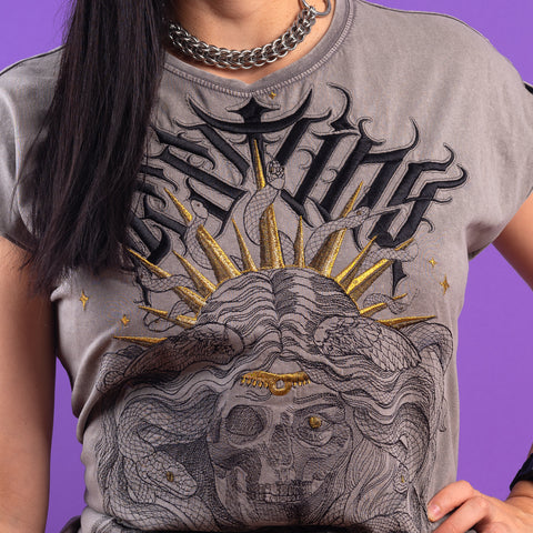 A person wears the Gorgone Embroidered Female T-shirt—a gray acid-wash tee with a skull, snakes, gold sun rays, and stylized text—paired with a chain necklace, standing against a purple background.