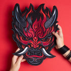 Someone holds the Oni Japanese Demon Embroidered Patch, featuring a fiery red and black demon mask with horns, sharp teeth, and intense eyes inspired by Japanese folklore, set against a matching red background.