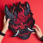 Two hands hold the Oni Japanese Demon Embroidered Patch, intricately designed with horns, sharp teeth, and tusks, inspired by Japanese folklore, set against a vibrant red background.