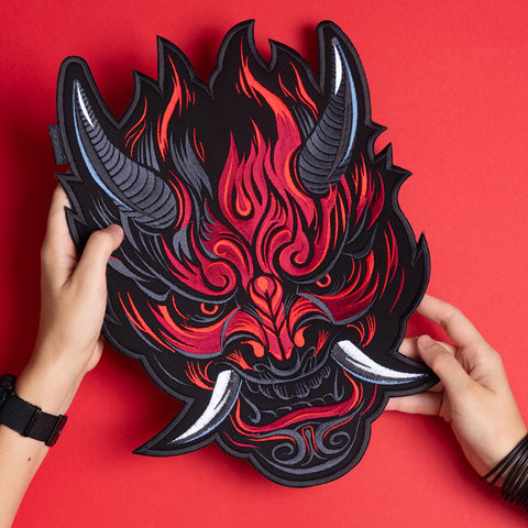Two hands hold the Oni Japanese Demon Embroidered Patch, intricately designed with horns, sharp teeth, and tusks, inspired by Japanese folklore, set against a vibrant red background.