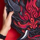 A hand holds the Oni Japanese Demon Embroidered Patch, showcasing a fierce red and black horned demon face with sharp teeth, inspired by Japanese folklore, set against a vibrant red background.
