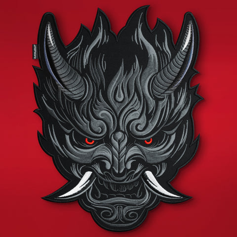 The Oni Japanese Demon Embroidered Patch features a stylized black and gray mask inspired by Japanese folklore, with sharp horns, tusks, glowing red eyes, flame-like patterns, and a fierce expression on a vivid red background.