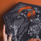 A hand displays the Raven and Skull Embroidered Back Patch: a black hexagonal patch with a detailed raven, a red-orange sun, white plants, and gothic motifs—ideal for fans of alternative fashion accessories.