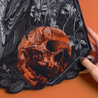 A hand holds the Raven and Skull Embroidered Back Patch, showcasing an orange skull with black shadows, gray floral and feather details, all set against an orange background.