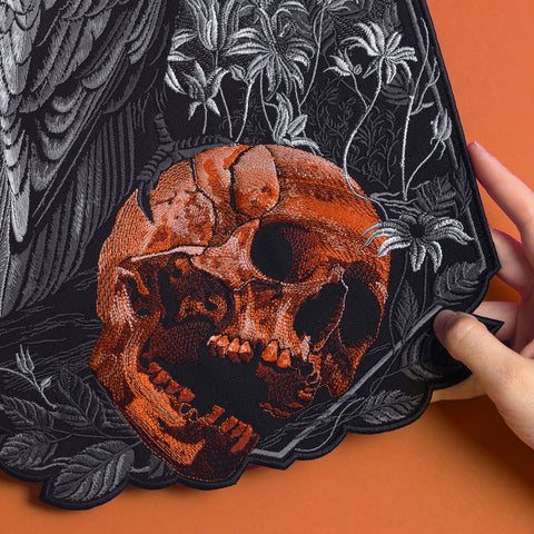 A hand holds the Raven and Skull Embroidered Back Patch, showcasing an orange skull with black shadows, gray floral and feather details, all set against an orange background.