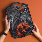 Two hands hold the Raven and Skull Embroidered Back Patch, featuring a black raven by a red-orange skull with plants and a red sun on an orange background—a bold gothic fashion accessory.