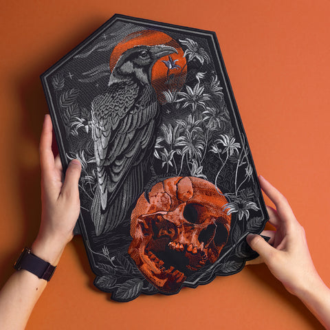Two hands hold the Raven and Skull Embroidered Back Patch, featuring a black raven by a red-orange skull with plants and a red sun on an orange background—a bold gothic fashion accessory.