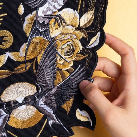 A hand holds the Spotify Song Code Embroidered Patch, featuring gold roses and detailed birds in flight on a light yellow background.