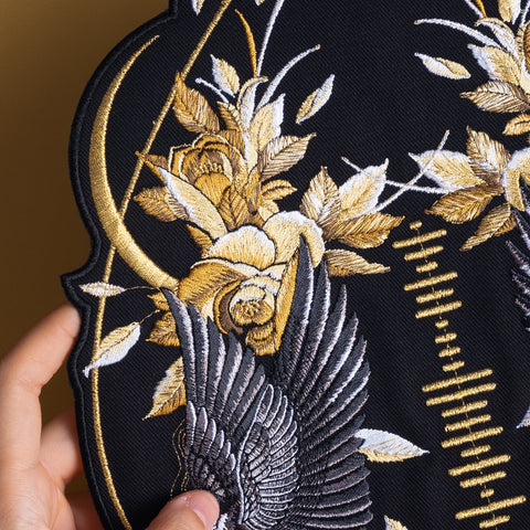 A hand holds a black Spotify Song Code Embroidered Patch with golden-yellow and white flowers, delicate leaves, a bird’s wing, and an intricate gold crescent and linear pattern in the background.