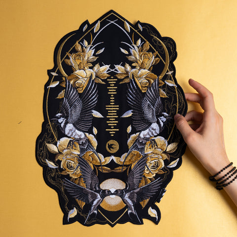 A hand holds a Spotify Song Code Embroidered Patch featuring mirrored birds, roses, leaves, and geometric patterns in gold, white, and gray on a black background, set against a yellow surface.