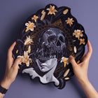 Two hands hold the Mass Hypnosis Embroidered Back Patch, featuring a gothic figure with a skull over their face, surrounded by yellow lilies and gold geometric designs on a purple background.