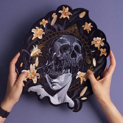Two hands hold the Mass Hypnosis Embroidered Back Patch, featuring a gothic figure with a skull over their face, surrounded by yellow lilies and gold geometric designs on a purple background.