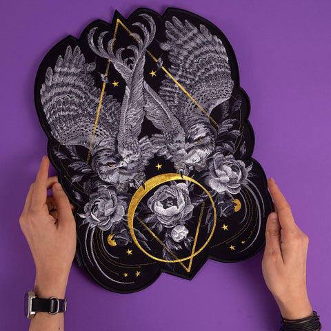 Two hands hold the Eagles Embroidered Custom Patch, showcasing detailed eagles, roses, and gold geometric accents on a purple background with celestial stars, swirling patterns, and intricate white and gold stitching.