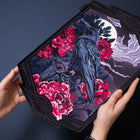 A person holds the Crow And Red Peonies Embroidered Patch, which features a black crow among vivid red peonies, with a full moon and dramatic clouds on an irregularly shaped dark fabric—a striking piece of Japanese-inspired art.