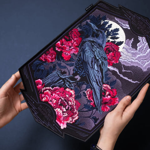 A person holds the Crow And Red Peonies Embroidered Patch, which features a black crow among vivid red peonies, with a full moon and dramatic clouds on an irregularly shaped dark fabric—a striking piece of Japanese-inspired art.