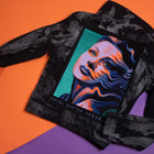 A black acid-wash denim jacket features the Vaporware Synth Pop Embroidered Back Patch: a vibrant cyberpunk design showing a woman's face distorted in waves with "FATAL SYSTEM ERROR," set against an orange and purple background.