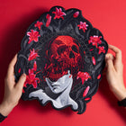 A person holds the Mass Hypnosis Embroidered Back Patch, featuring a red skull over a grayscale face with red lilies on a black background, set against a vibrant red backdrop.