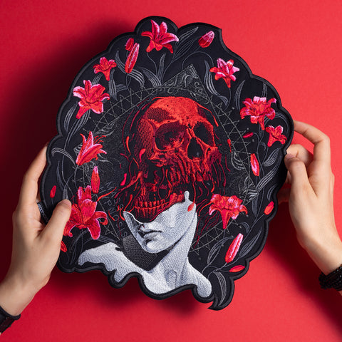 A person holds the Mass Hypnosis Embroidered Back Patch, featuring a red skull over a grayscale face with red lilies on a black background, set against a vibrant red backdrop.