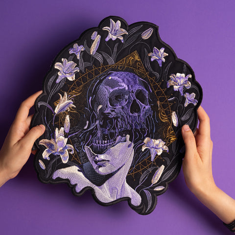 Two hands hold the Mass Hypnosis Embroidered Back Patch, showcasing a skull merging with a woman’s face, white lilies, and gold geometric lines on a purple background.