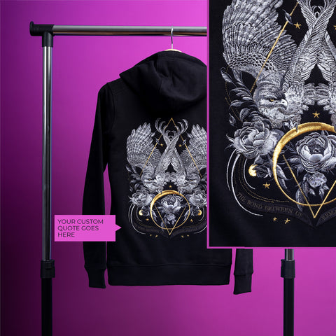 The Eagles Unisex Embroidered Hoodie, in black, features an intricate silver and gold design of two birds, roses, and stars on the back. Displayed on a rack with a bright purple backdrop and a pink tag that reads “Your custom quote goes here.”.