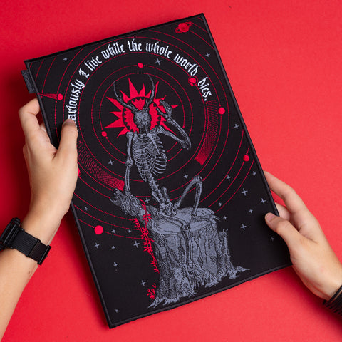 A person holds the Vicarious Embroidered Back Patch, featuring a skeleton on a tree stump, red rings, and "I live while the whole world dies" on a solid red background—ideal for fans of occult fashion.