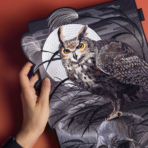 A hand holds the Owl & Skull Embroidered Back Patch, showing a detailed owl with orange eyes perched on a skull before a full moon and dark branches on a reddish-brown background, capturing pastel goth style.