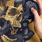 A hand holds a detailed "Snake Skeleton Embroidered Patch" featuring a gold and gray skull with horns and roses on a black background.