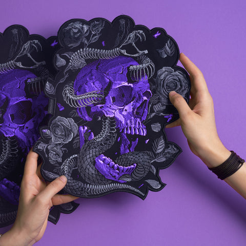 Two hands hold the Snake Skeleton Embroidered Patch, showcasing a purple and black skull with roses and a snake, set against a matching purple background.