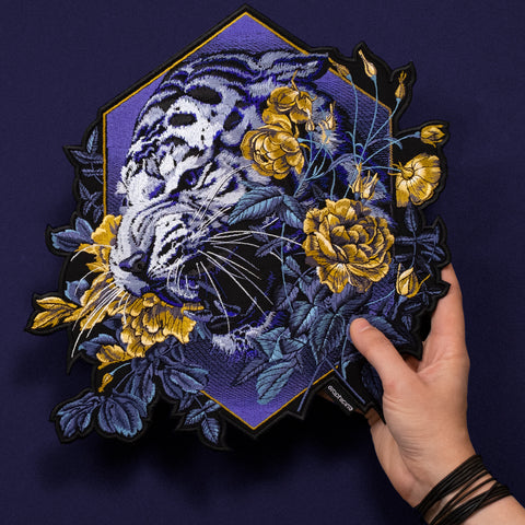 A hand holds the Tiger and Roses Embroidered Patch, showing a white tiger among yellow flowers and blue leaves on a dark blue background. The hexagonal border and detailed stitching give it a unique bohemian style.