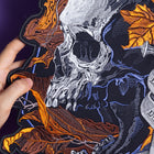 A hand holds the Skull and Dagger Embroidered Back Patch, showcasing a detailed white skull, orange autumn leaves, a sword, and a "DEATH BEFORE DISHONOR" banner—perfect for gothic fashion—set on a dark background.