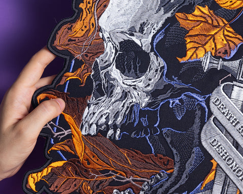 A hand holds the Skull and Dagger Embroidered Back Patch, showcasing a detailed white skull, orange autumn leaves, a sword, and a "DEATH BEFORE DISHONOR" banner—perfect for gothic fashion—set on a dark background.