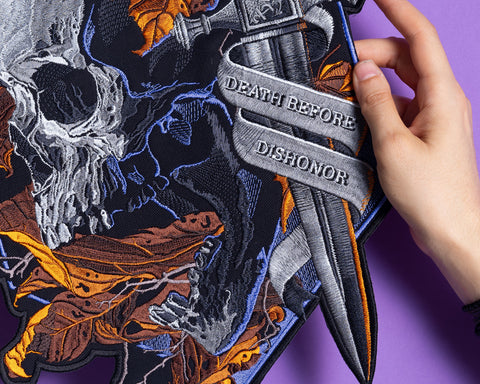 A hand displays the Skull and Dagger Embroidered Back Patch, featuring autumn leaves and a dagger with "DEATH BEFORE DISHONOR," ideal for gothic fashion fans, set against a purple background.