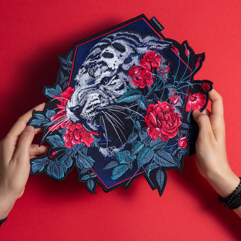 Two hands hold the Tiger and Roses Embroidered Patch, showcasing a roaring white tiger, red roses, and green leaves—a bold boho accessory with a vivid red background, perfect for adding unique flair to your style.
