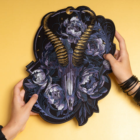 Two hands hold the Blesbok Skull and Flowers Embroidered Patch, featuring a gold-horned ram skull, purple flowers, and dark leaves on yellow—perfect for goth fashion enthusiasts.