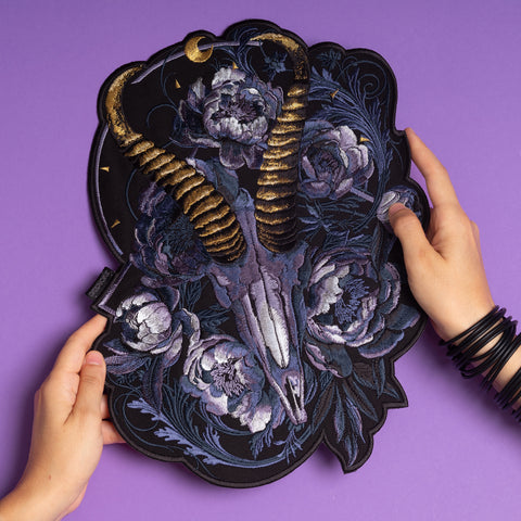 Two hands hold the Blesbok Skull and Flowers Embroidered Patch, featuring a golden-horned ram skull with purple flowers and leaves—a bold back patch for goth fashion, beautifully displayed on a purple background.