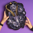 Two hands hold the Blesbok Skull and Flowers Embroidered Patch, featuring a ram skull with gold horns, white flowers, and dark foliage—an eye-catching back patch for goth fashion, displayed on a purple background.