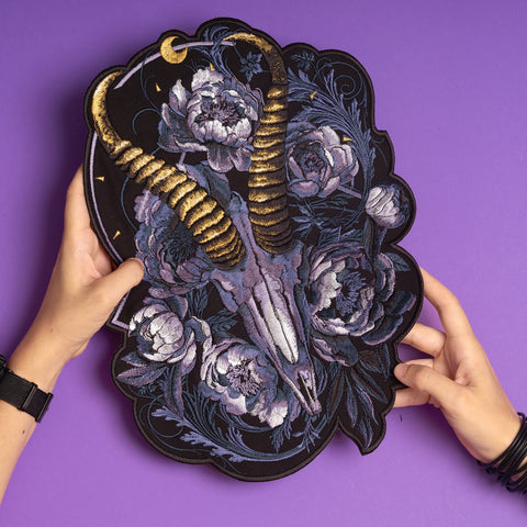 Two hands hold the Blesbok Skull and Flowers Embroidered Patch, featuring a ram skull with gold horns, white flowers, and dark foliage—an eye-catching back patch for goth fashion, displayed on a purple background.