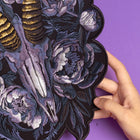A hand holds the Blesbok Skull and Flowers Embroidered Patch, featuring a detailed animal skull with gold horns and purple and white floral accents—an ideal accessory to add gothic flair to your style.