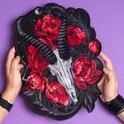 A person holds the Blesbok Skull and Flowers Embroidered Patch, featuring a goat skull with black horns, vivid red peonies, and dark leaves on black. Black bracelets adorn their hands, set against a bold purple background—ideal for goth style.