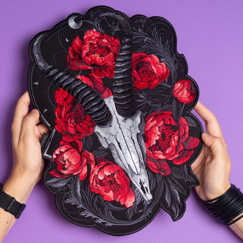 A person holds the Blesbok Skull and Flowers Embroidered Patch, featuring a goat skull with black horns, vivid red peonies, and dark leaves on black. Black bracelets adorn their hands, set against a bold purple background—ideal for goth style.