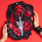 A person holds the Blesbok Skull and Flowers Embroidered Patch, featuring a silver ram skull with black horns, red flowers, and dark leaves on a red background, styled with black bangles for a bold goth fashion look.