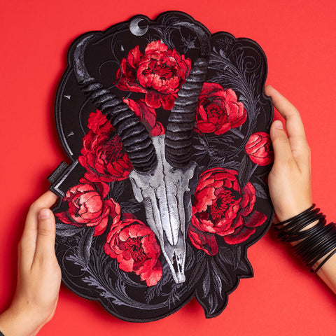 A person holds the Blesbok Skull and Flowers Embroidered Patch, featuring a silver ram skull with black horns, red flowers, and dark leaves on a red background, styled with black bangles for a bold goth fashion look.