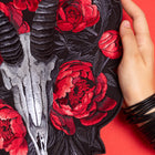 A hand holds the Blesbok Skull and Flowers Embroidered Patch, showcasing a silver ram skull with black horns and vivid red flowers—an ideal accessory for goth fashion fans.